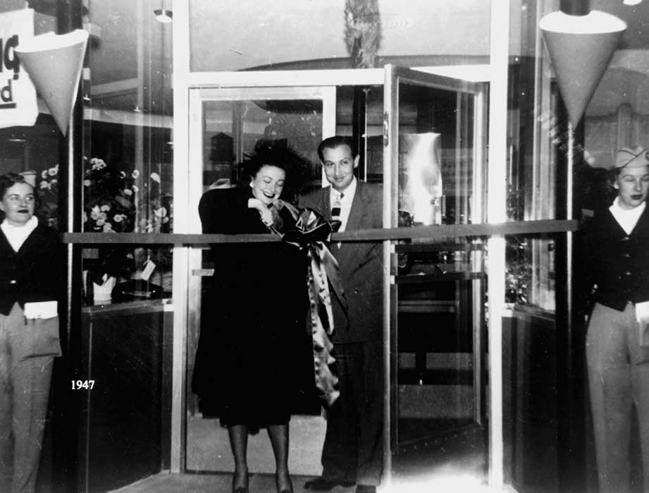 A woman cuts a ribbon at a store entrance, possibly Mel's Drive-In Restaurant, while a man smiles beside her. Two women in uniforms watch from each end. The scene appears to be from 1947, as noted in the image's corner—an iconic diner moment.
