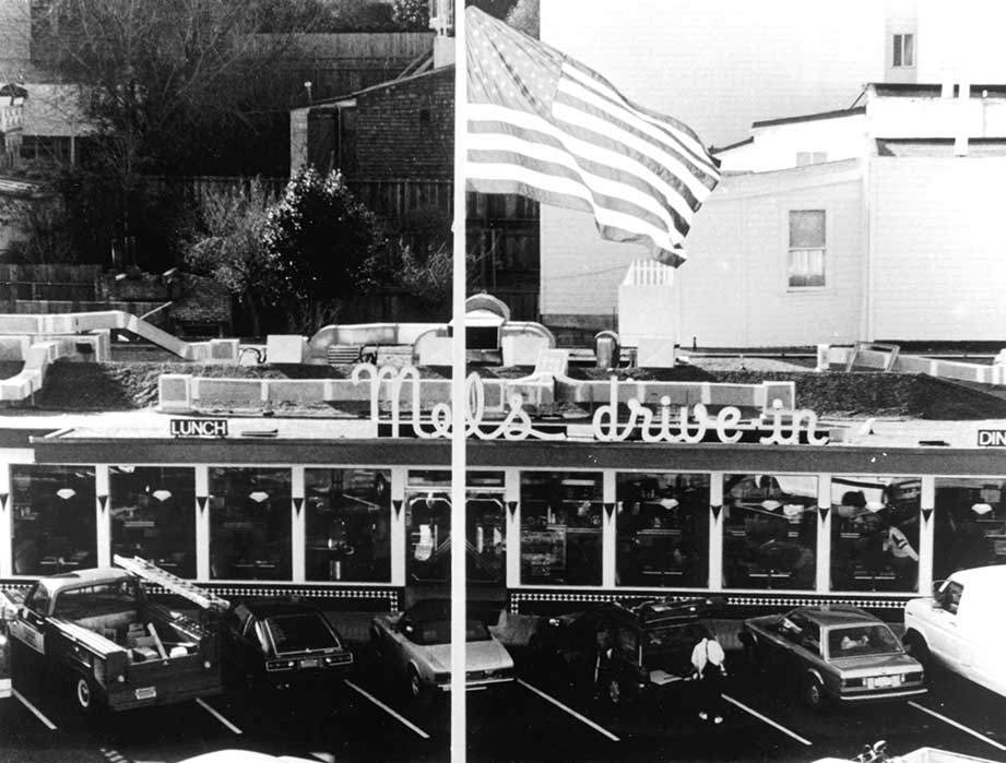 Black and white photo of the iconic diner, Mel's Drive-In Restaurant, with a large American flag in front, several parked cars in the lot, and people visible inside. The neon sign reads Mels drive-in.