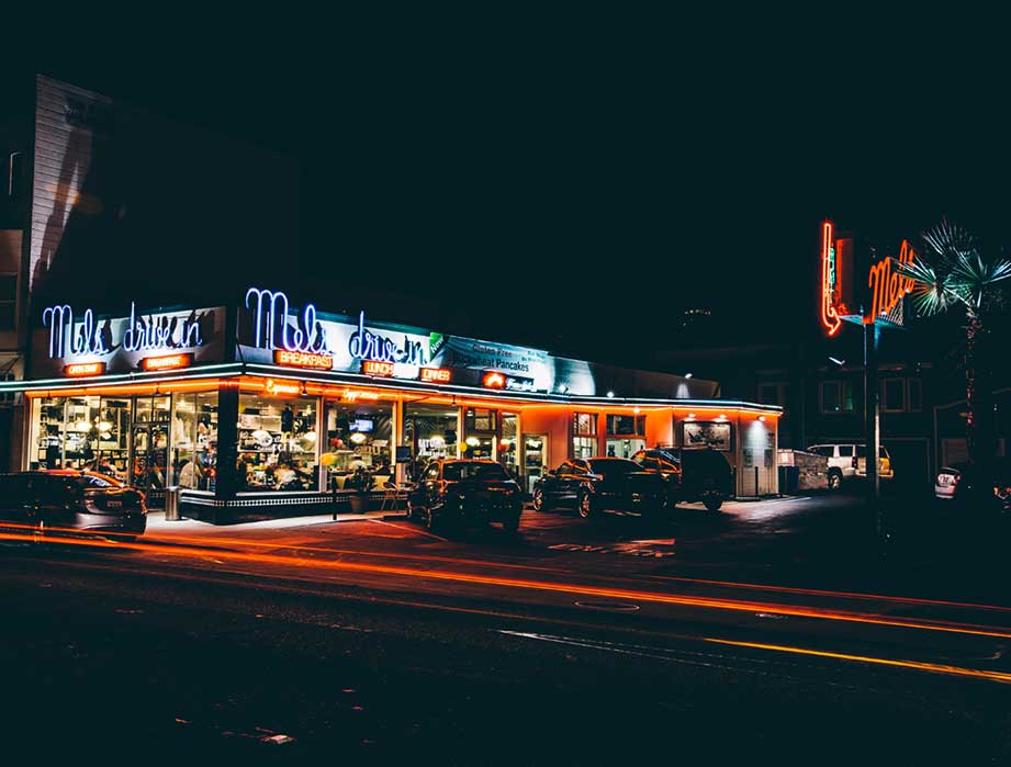 A retro-style night scene at the iconic Mel’s Drive-In Restaurant, brightly lit with neon signs. Several cars are parked outside, and light trails from passing vehicles illuminate this classic diner atmosphere.