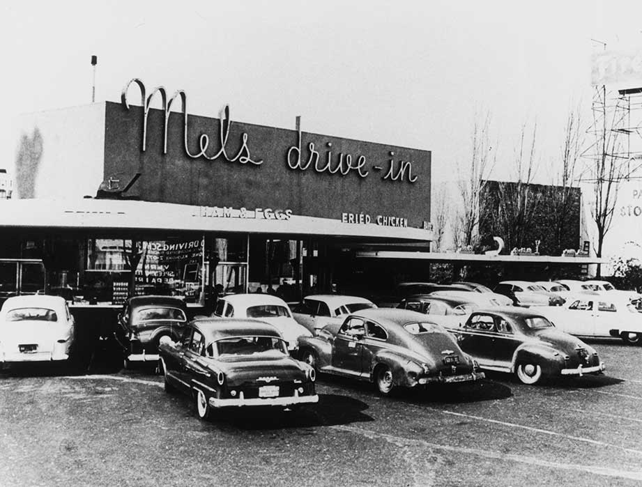 Black-and-white photo of vintage cars parked outside the iconic diner, Mel’s Drive-In Restaurant. The building has large neon signage, and people can be seen inside and around the retro-style eatery.