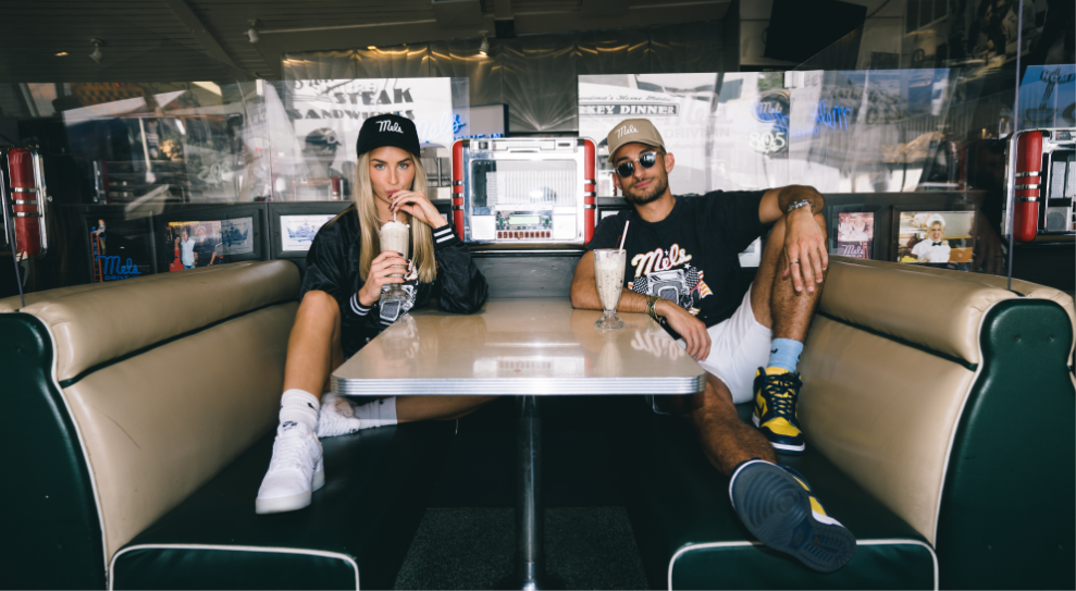 Two people sit casually in an iconic diner booth at Mel's Drive-In Restaurant. One sips a milkshake, both in streetwear and caps, while a jukebox sits on the table. The overall vibe is relaxed and stylish.