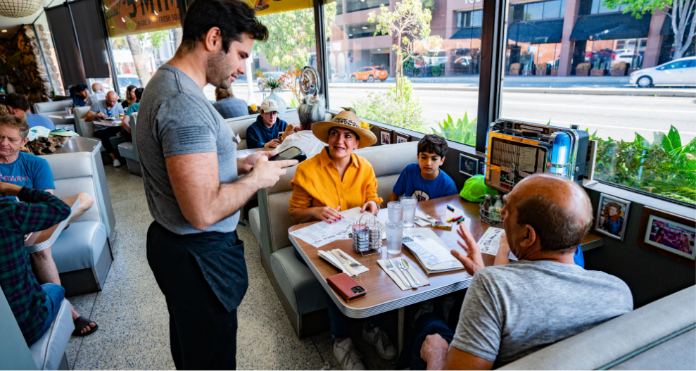 A waiter takes an order from three people at a booth in the iconic Mel's Drive-In Restaurant, its large windows flooding the diner with light as a woman in a hat, a young boy, and an older man sit together facing the street.