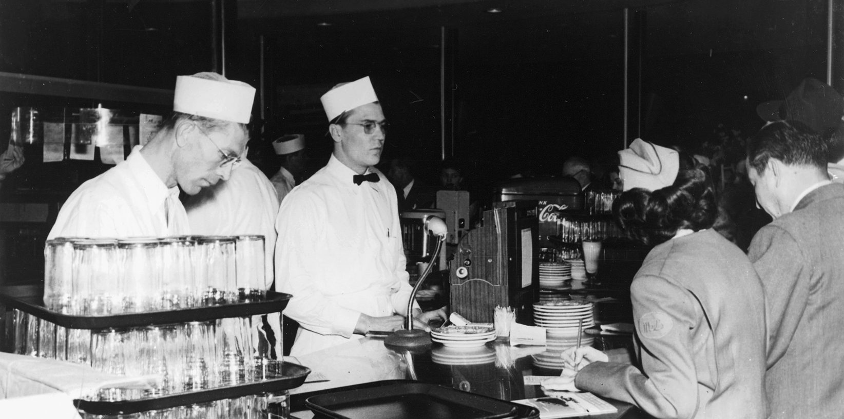 Black and white photo of male soda fountain workers in white uniforms and hats serving customers at the busy Mel's Drive-In Restaurant counter, with stacked glasses and plates visible—an iconic diner scene.