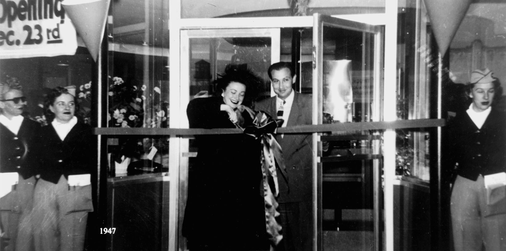 Black and white photo of a woman cutting a large ribbon at a doorway, with a man beside her. Three women in uniforms stand nearby. A sign reads Opening Dec. 23rd. The year 1947 is marked in the corner—an iconic diner moment at Mel's Drive-In Restaurant.