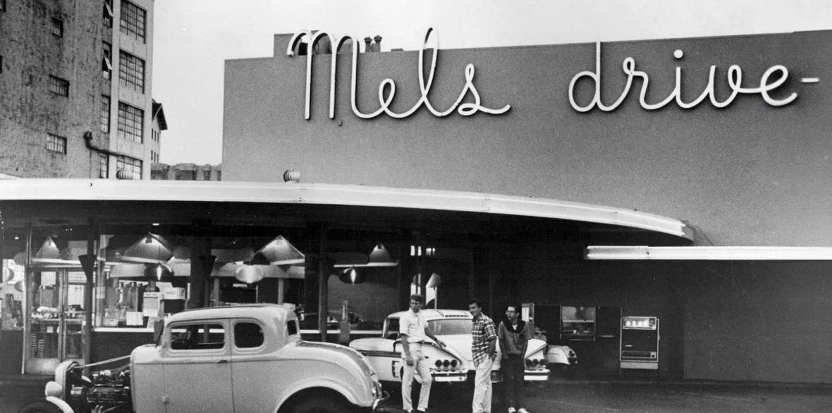 Black-and-white photo of Mel’s Drive-In Restaurant, an iconic diner with classic cars parked out front and three people standing near the entrance under a large neon sign that reads “Mel’s drive-.”.