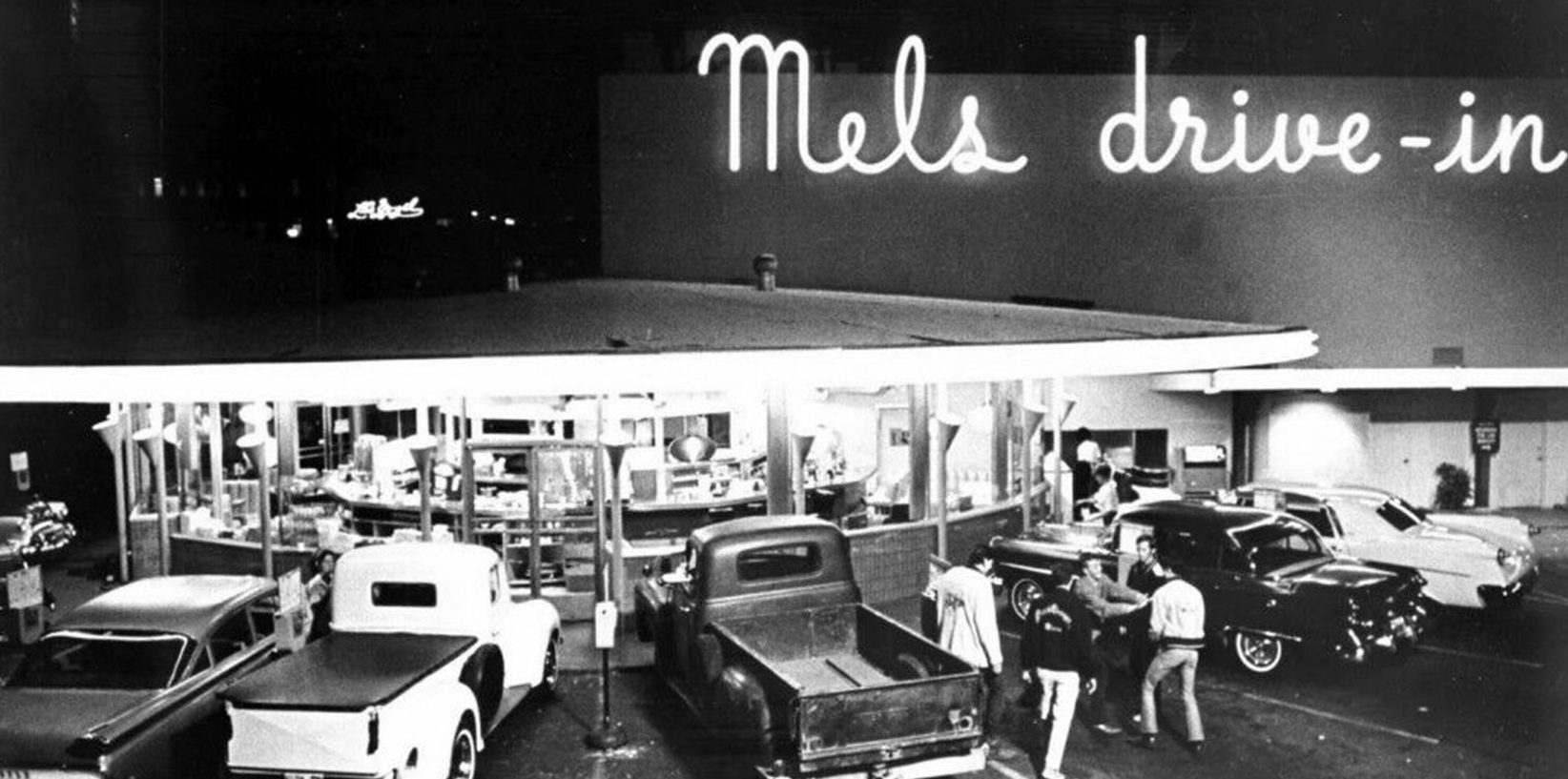 A black-and-white photo of the iconic diner Mel's Drive-In Restaurant at night, with a neon sign glowing above vintage cars and people gathered near the entrance.