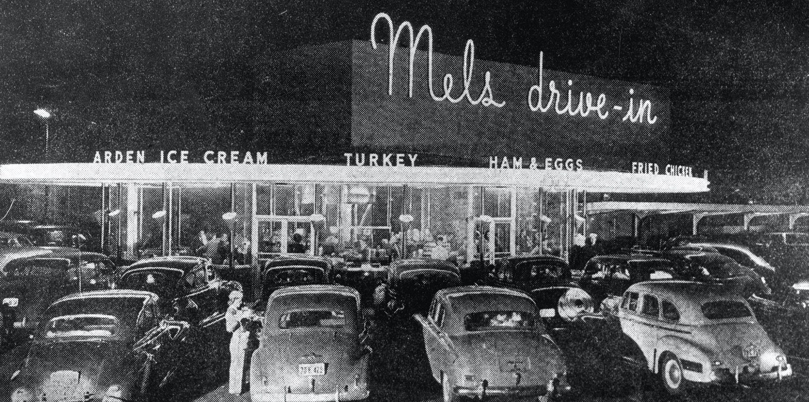 Black-and-white photo of the iconic diner, Mel’s Drive-In Restaurant, at night. A neon sign glows above a crowded lot of vintage cars as people socialize near the entrance and diners gather inside the brightly lit restaurant.