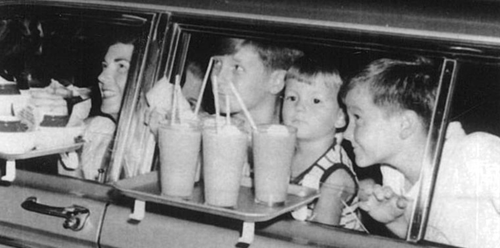 A black-and-white photo shows four children inside a car at a drive-in, looking out the window. A tray with drinks and food from Mel's Drive-In Restaurant, an iconic diner, is attached to the car door outside their window.
