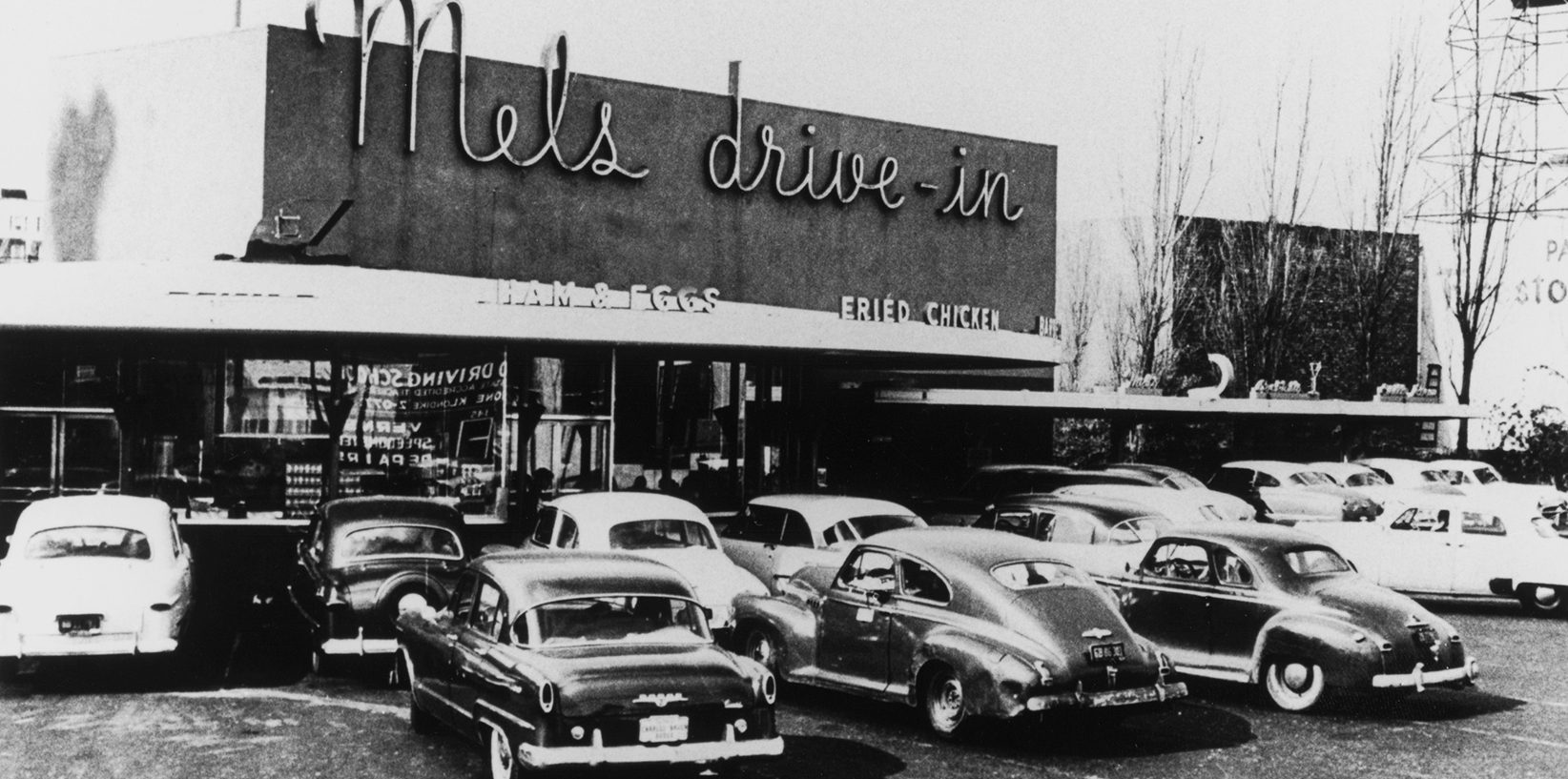 Black-and-white photo of classic cars parked in front of Mel’s Drive-In Restaurant, an iconic diner with a large neon sign, glass windows, and outdoor signs advertising ham, eggs, and fried chicken.