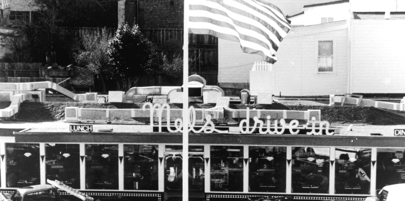 Black and white photo of a vintage diner with a neon sign reading Mels drive-in. An American flag waves above the building, and cars are visible in the background. The diner has large windows and retro decor.